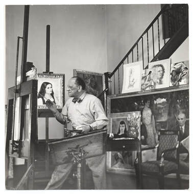 Photo of Leon Kroll painting in his studio, surrounded by works of art.